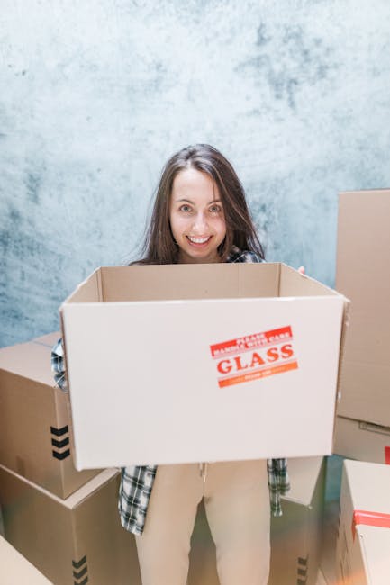 A man with dark curly hair and a beard, dressed in a blue t-shirt, stands inside a room during a home relocation process, with his hands resting on a large cardboard box. The room has white walls, a w